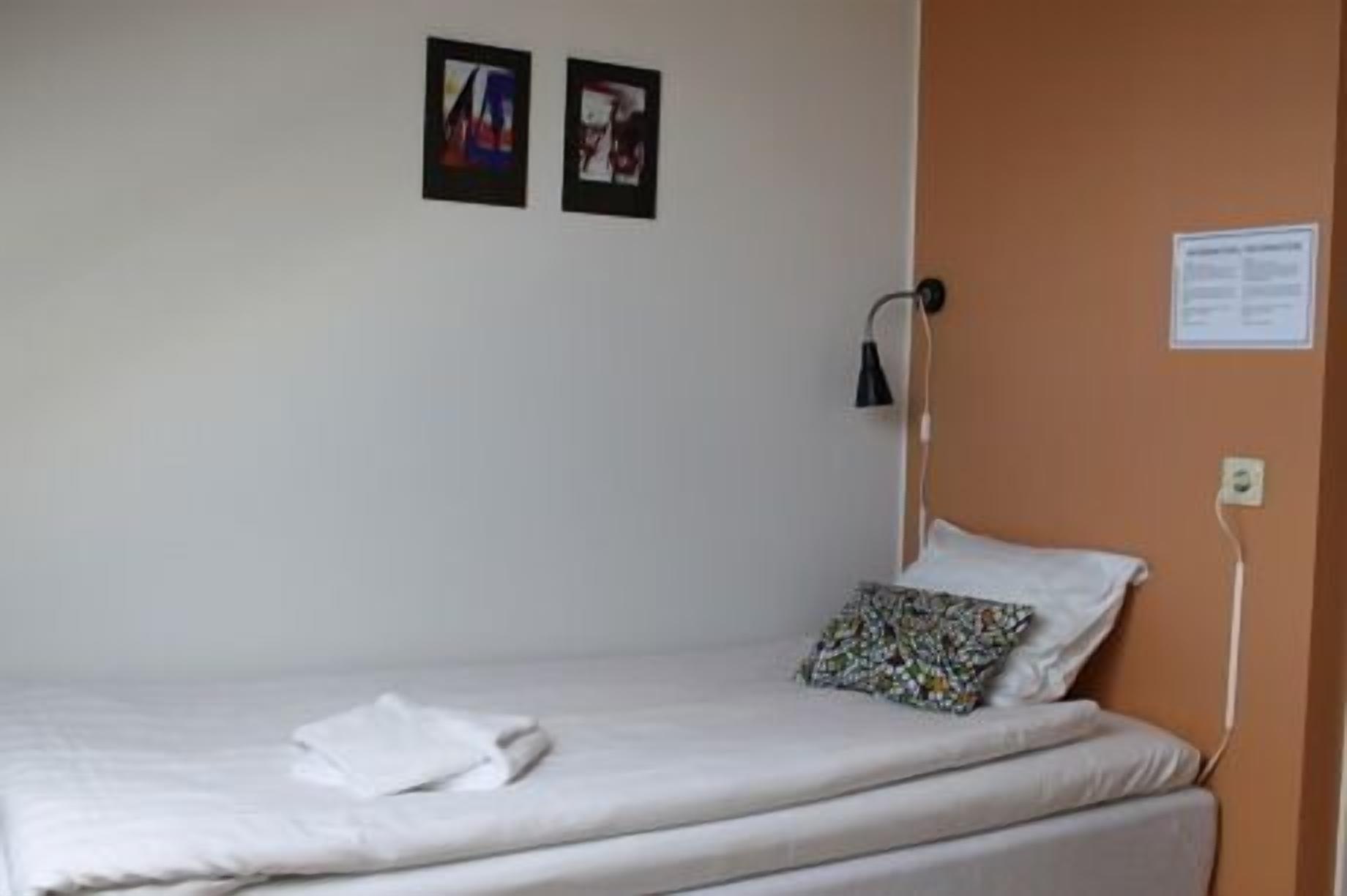 Ein Einzelbett mit weißer Bettwäsche, einem gemusterten Kissen und einer Wandlampe. Zwei gerahmte Bilder hängen an der Wand über dem Bett.