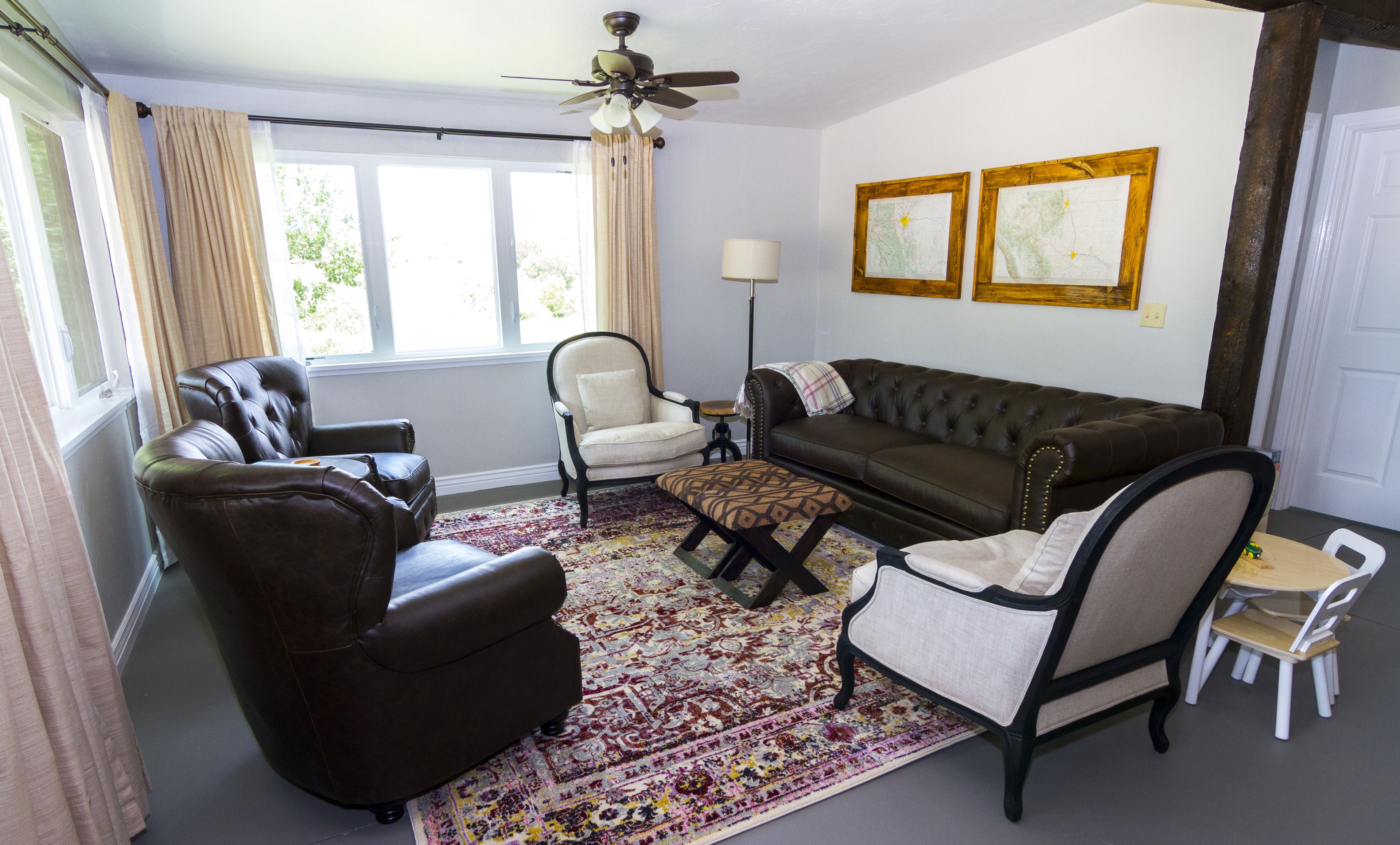 Ein Wohnzimmer mit einem braunen Ledersofa, zwei Sesseln, einem gemusterten Teppich, einem Deckenventilator und zwei gerahmten Karten an der Wand.