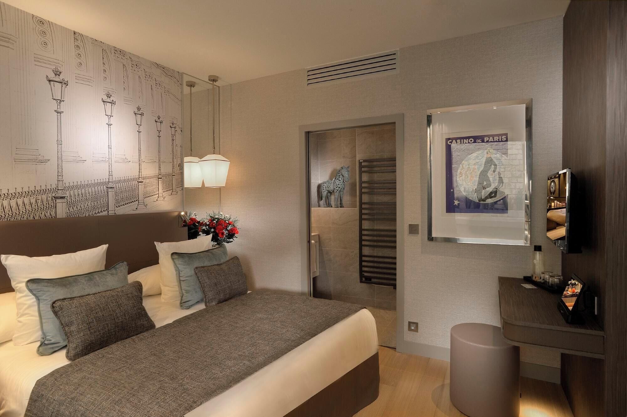 Una habitación de hotel moderna con una cama grande, una mesita de noche, un televisor montado en la pared y un cartel del Casino de París.