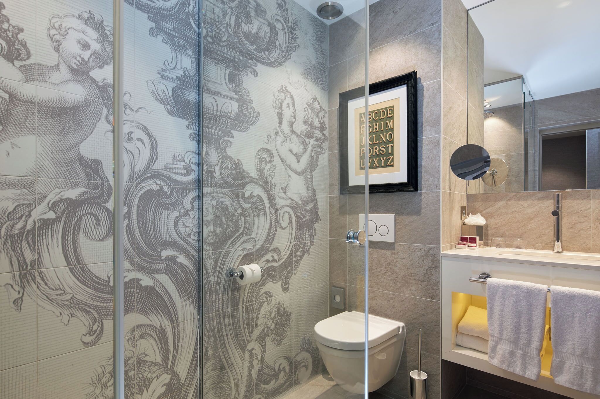 Een badkamer met douche, toilet en wastafel. Aan de muur hangt een alfabetkaart in een lijst en een handdoekrek met handdoeken.
