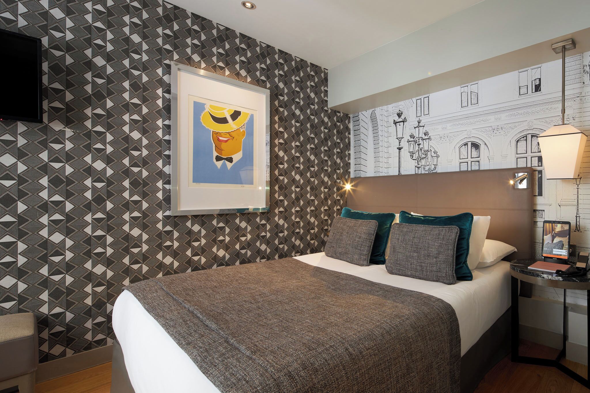 A hotel room with a patterned wallpaper, a framed picture on the wall, a bed with pillows, a bedside table with a lamp, and a TV.