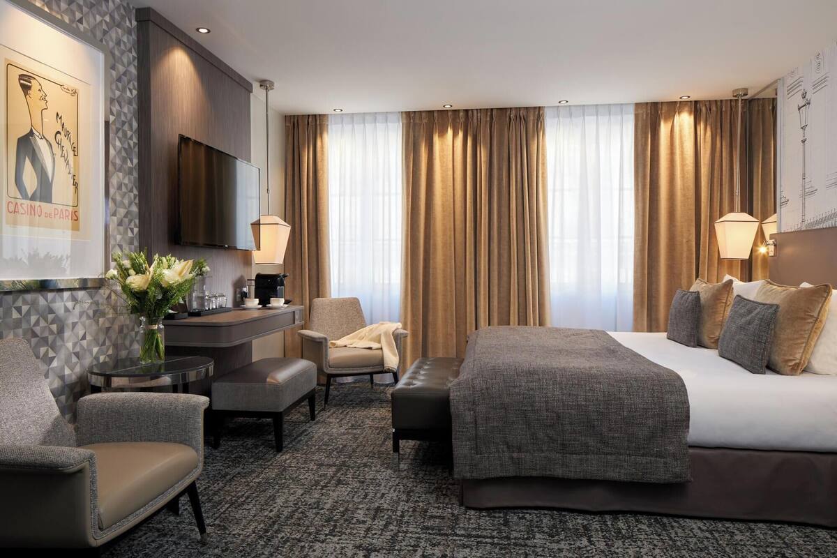 A hotel room with a bed, two armchairs, a desk with a lamp, a coffee machine, and a painting on the wall.