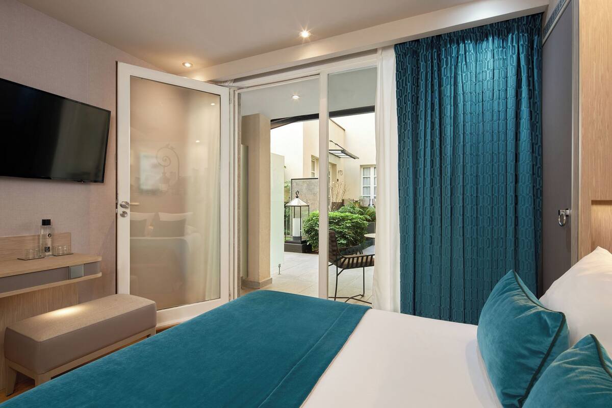 A modern hotel room with a large bed, a TV mounted on the wall, a bench, and a sliding glass door leading to an outdoor area.