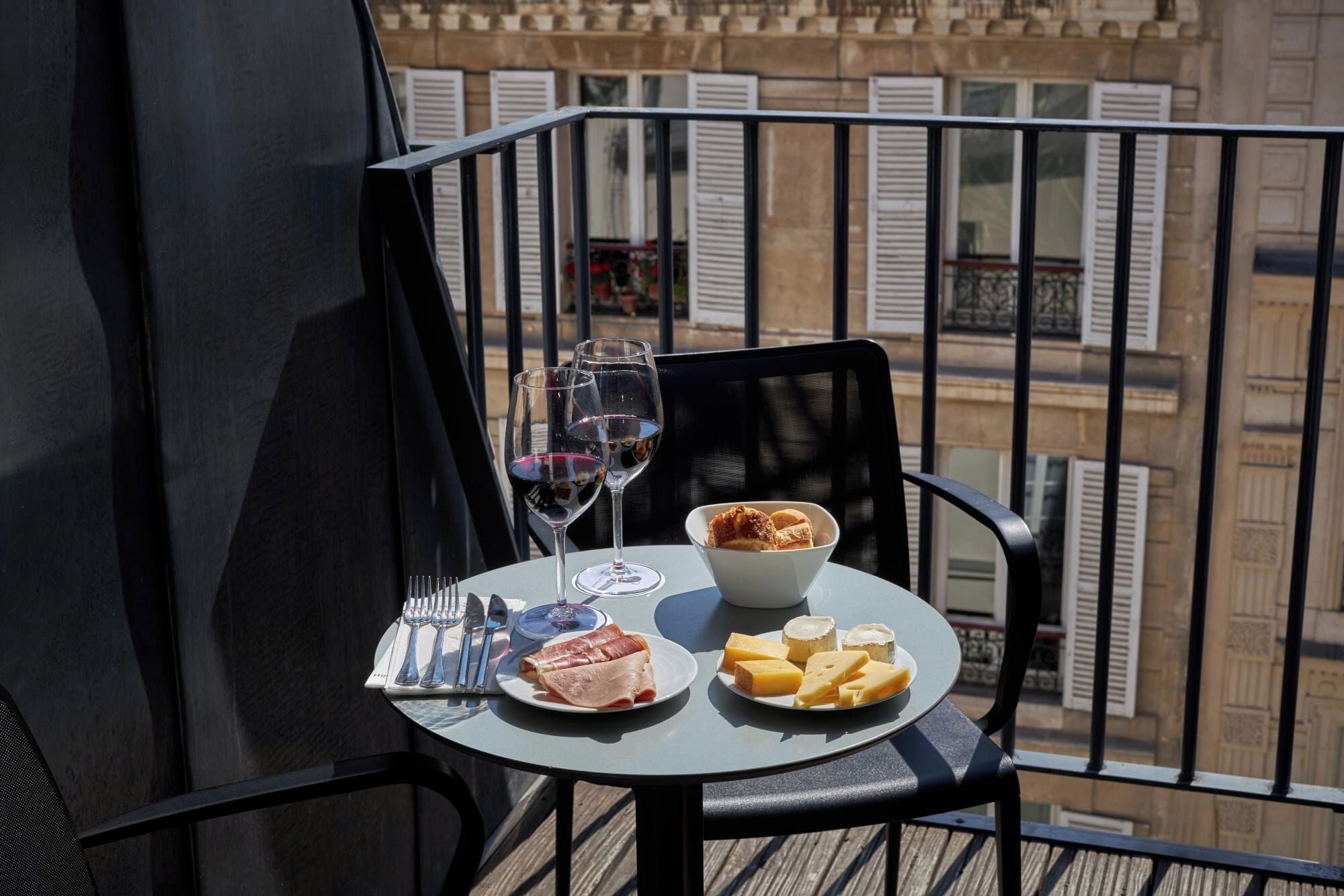 A balcony table with a meal setup including plates of food, glasses of wine, and a bowl of pastries.