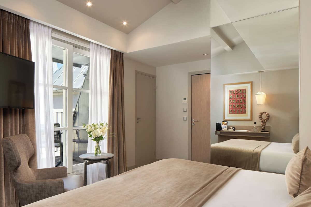 A hotel room with a bed, a chair, a small table with a vase, a mirror, and a window with curtains.