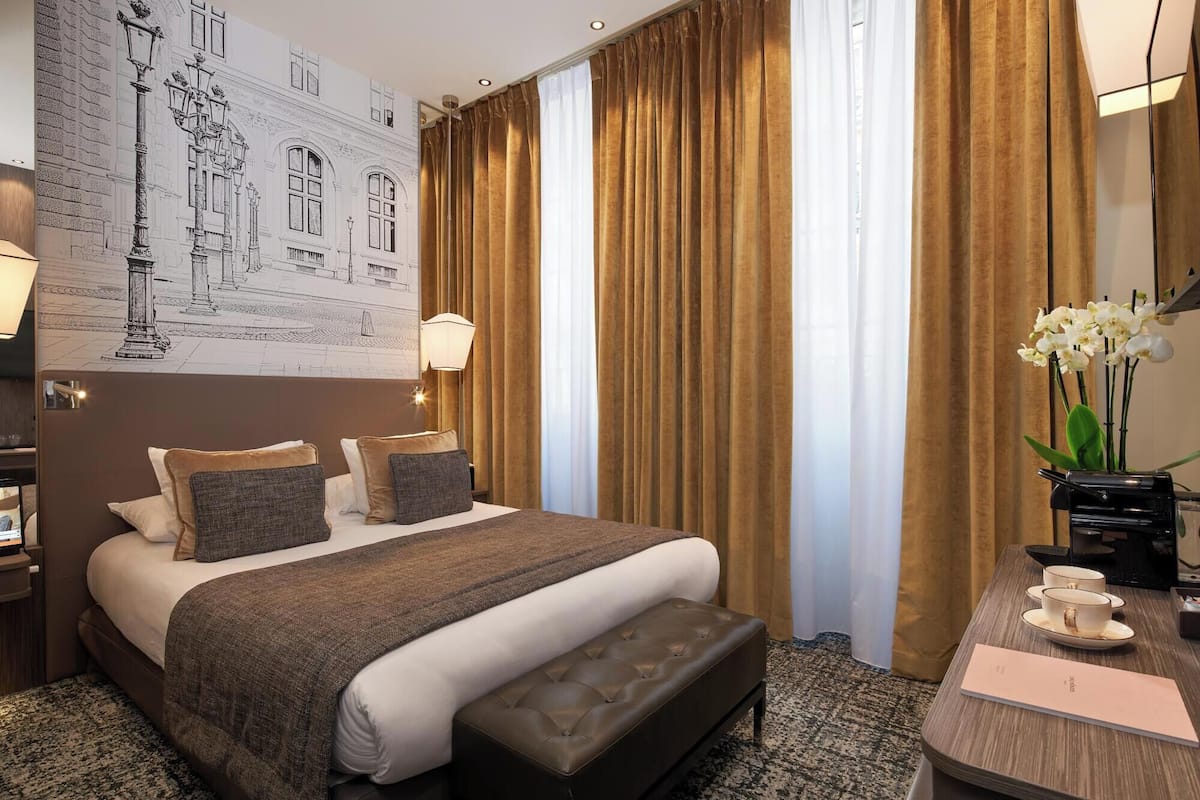 A hotel room with a large bed, a desk with a chair, a nightstand, and a wall-mounted lamp.