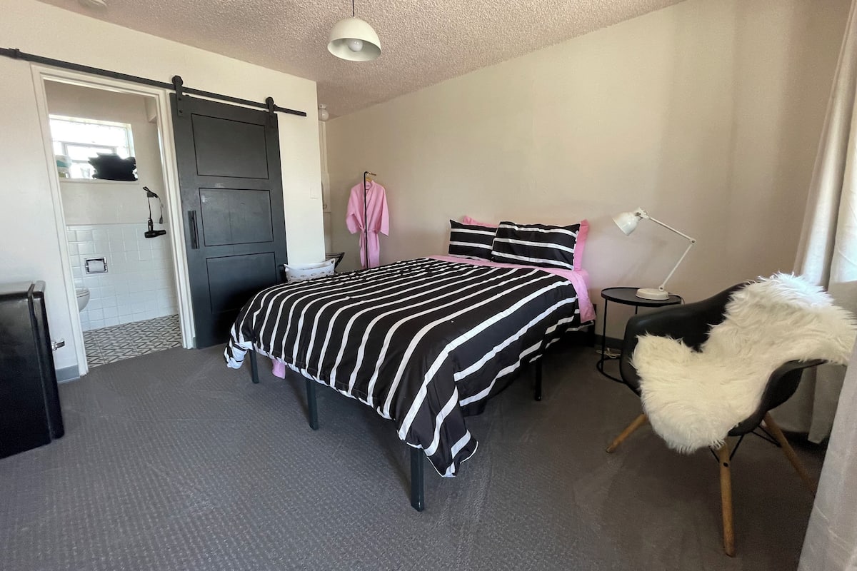 A bedroom with a bed, a bedside lamp, a chair with a fur throw, a robe hanging on the wall, and a door leading to a bathroom.