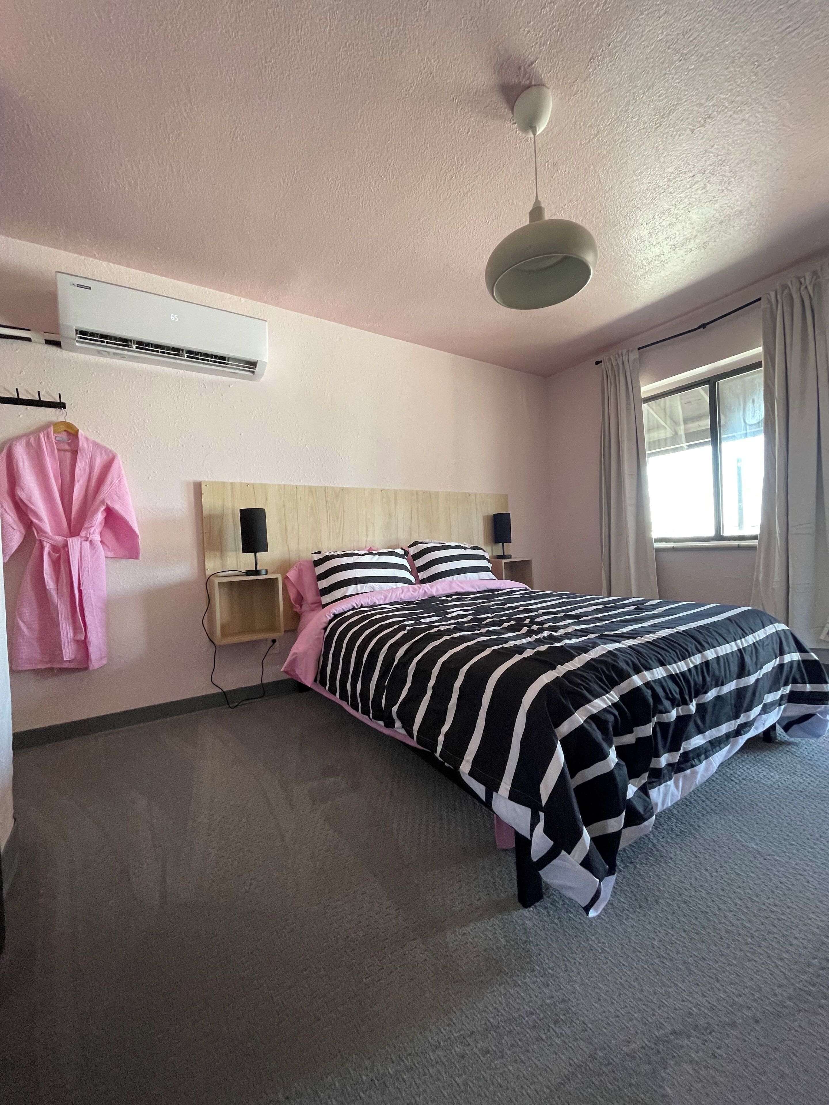 A hotel room with a bed, a bedside table with a lamp, a hanging robe, and an air conditioning unit.