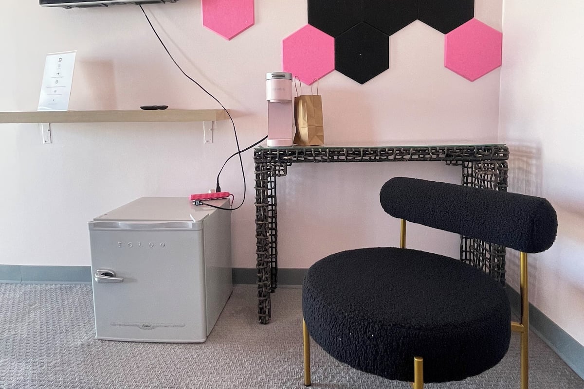 A room with a flat-screen TV mounted on the wall, a grey storage unit, a coffee maker on a table, and a black upholstered chair with gold legs.