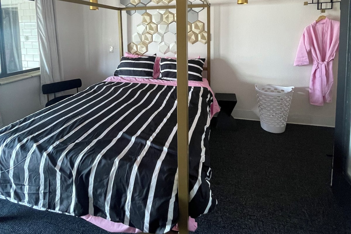 A four-poster bed with a geometric headboard, a window with curtains, a hanging light fixture, a wall-mounted towel rack, and a trash bin.