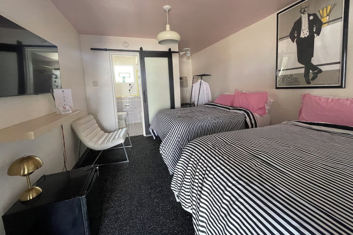 A hotel room with two beds, a desk, a chair, a mirror, a hanging light fixture, and a framed picture on the wall.