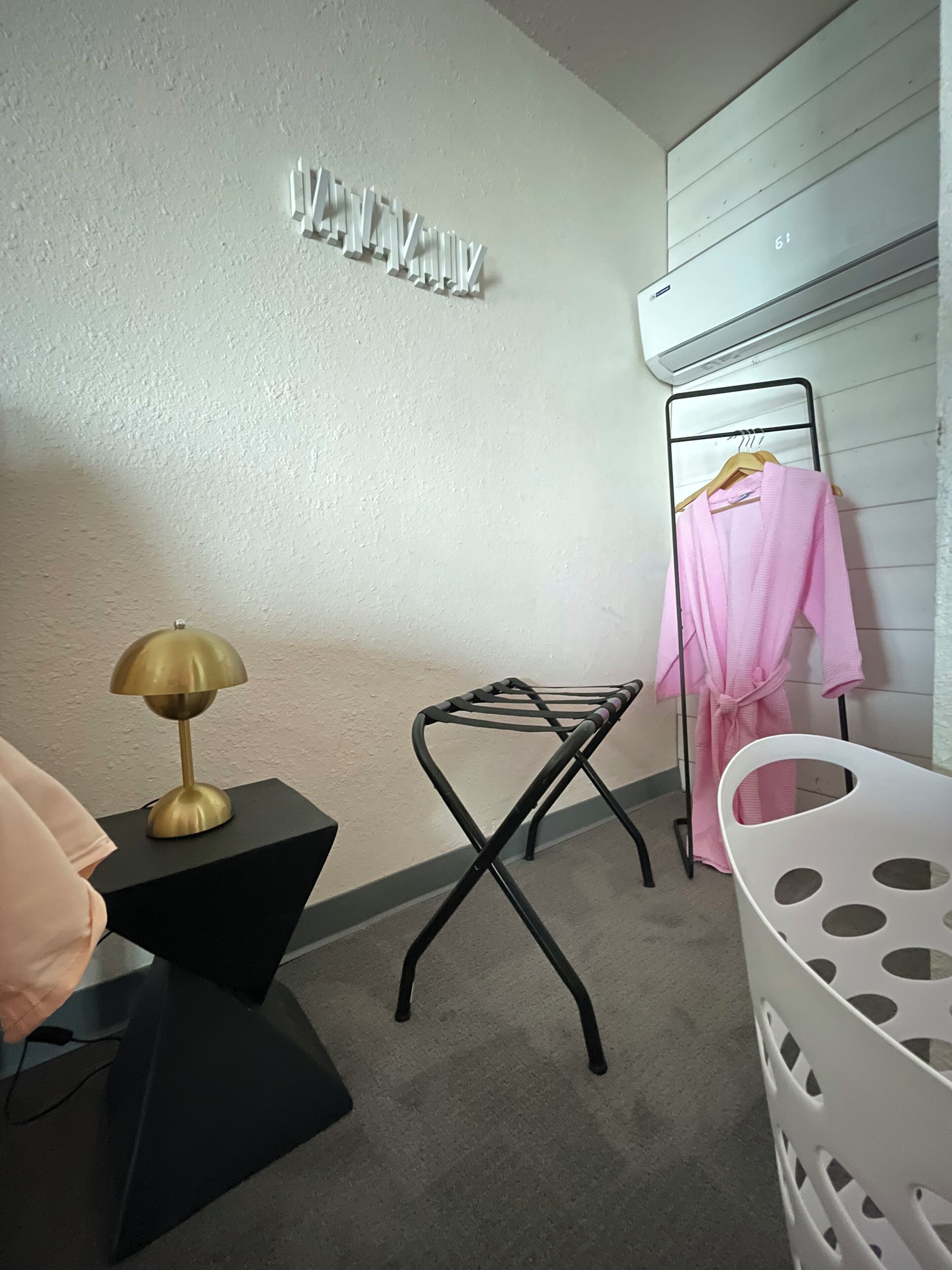 Una habitación con una mesita de noche negra, una lámpara dorada, una silla plegable negra, un cesto de ropa blanca y un tendedero de pared con ropa.