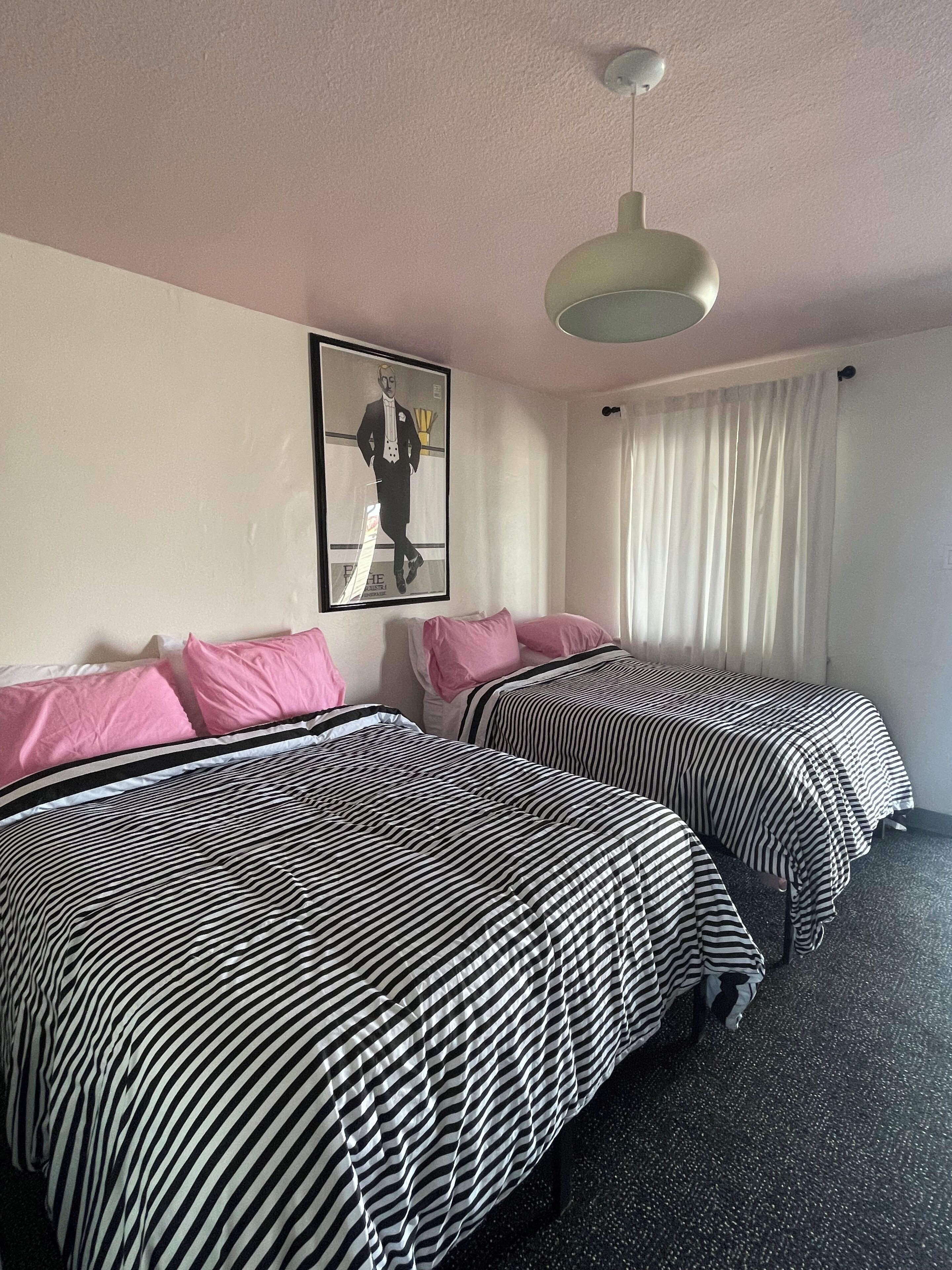 Hotelkamer met twee bedden, elk met streepjesbeddengoed, een plafondlamp, een ingelijst schilderij aan de muur en een raam met gordijnen.