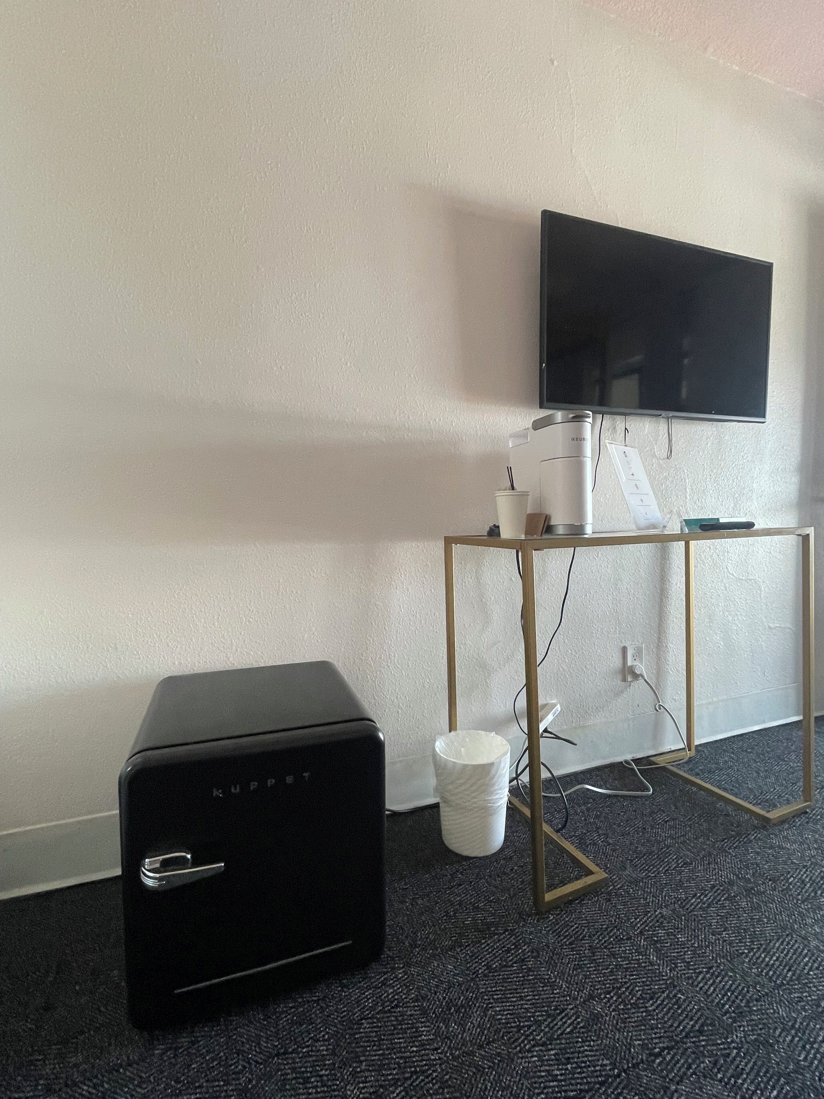 Une pièce comprenant un radiateur noir mobile, une poubelle blanche, une petite table avec une bouilloire et une tasse, et un téléviseur fixé au mur.