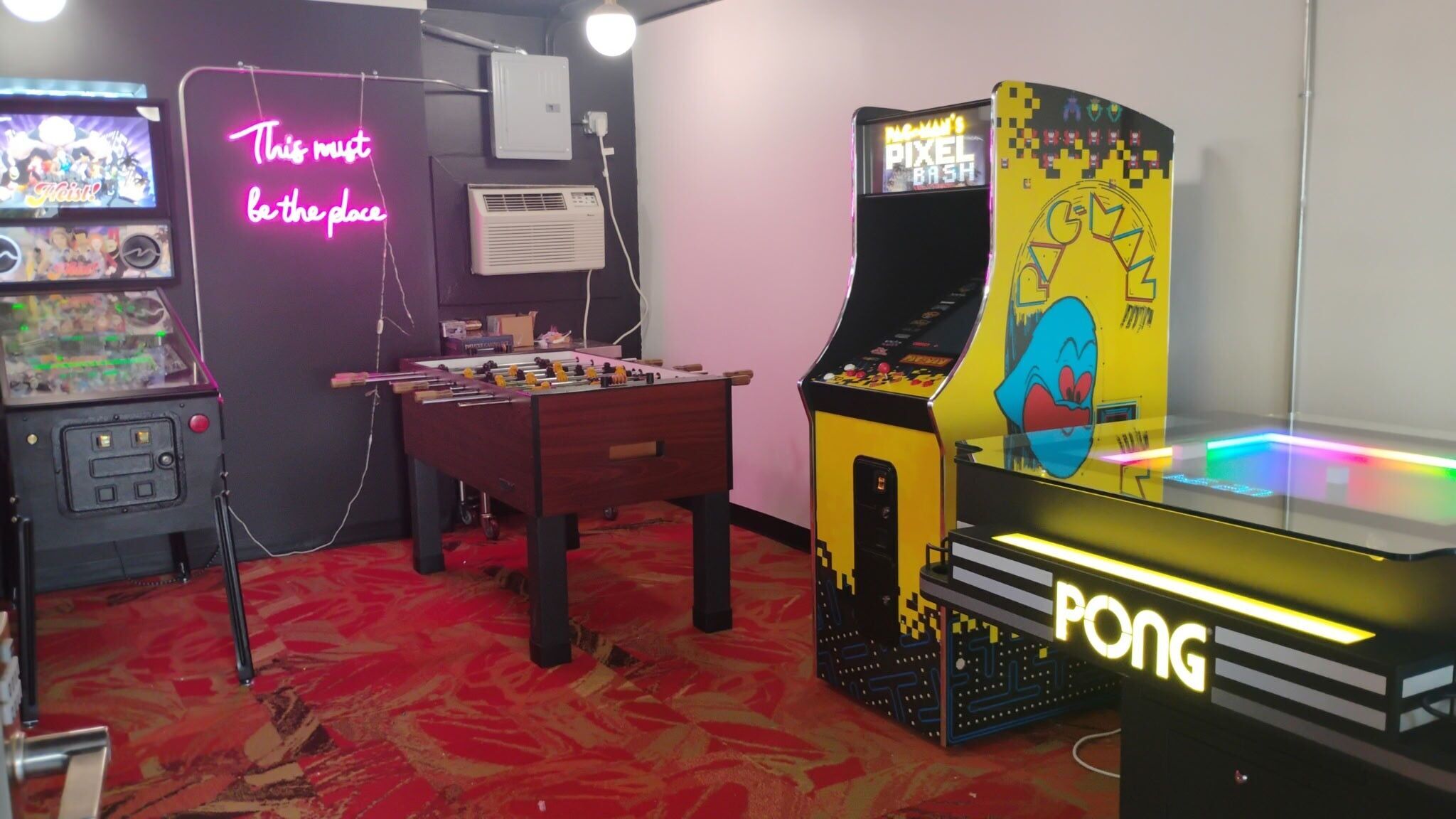 A room with a foosball table, a Pac-Man arcade machine, and a neon sign.