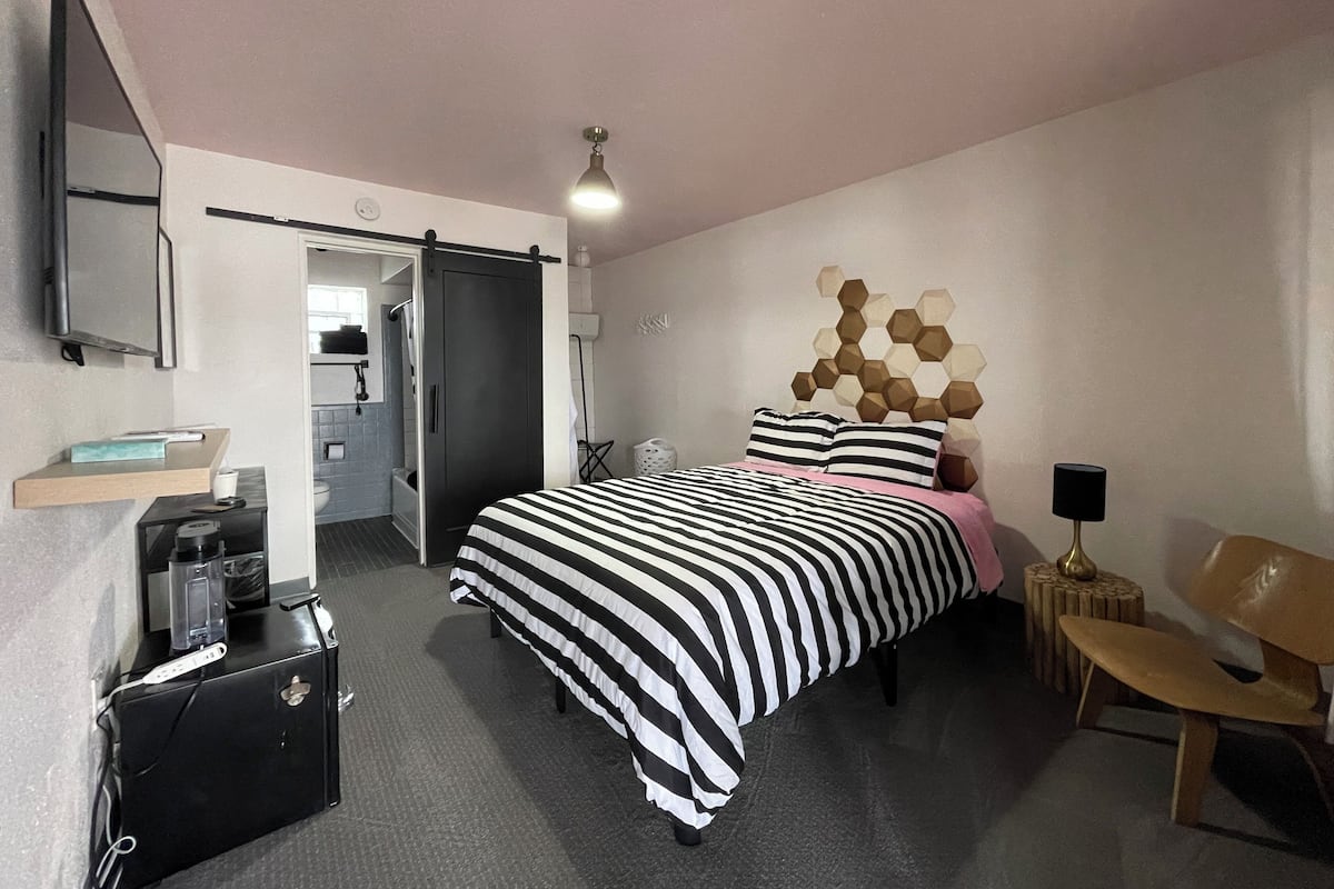 A modern bedroom with a large bed, a desk, a chair, a wardrobe, and a bathroom visible through an open door.