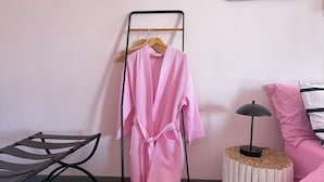 Een kamer met een bed, een houten nachtkastje, een kleine ronde tafel met een lamp en een metalen rek met een roze badjas.