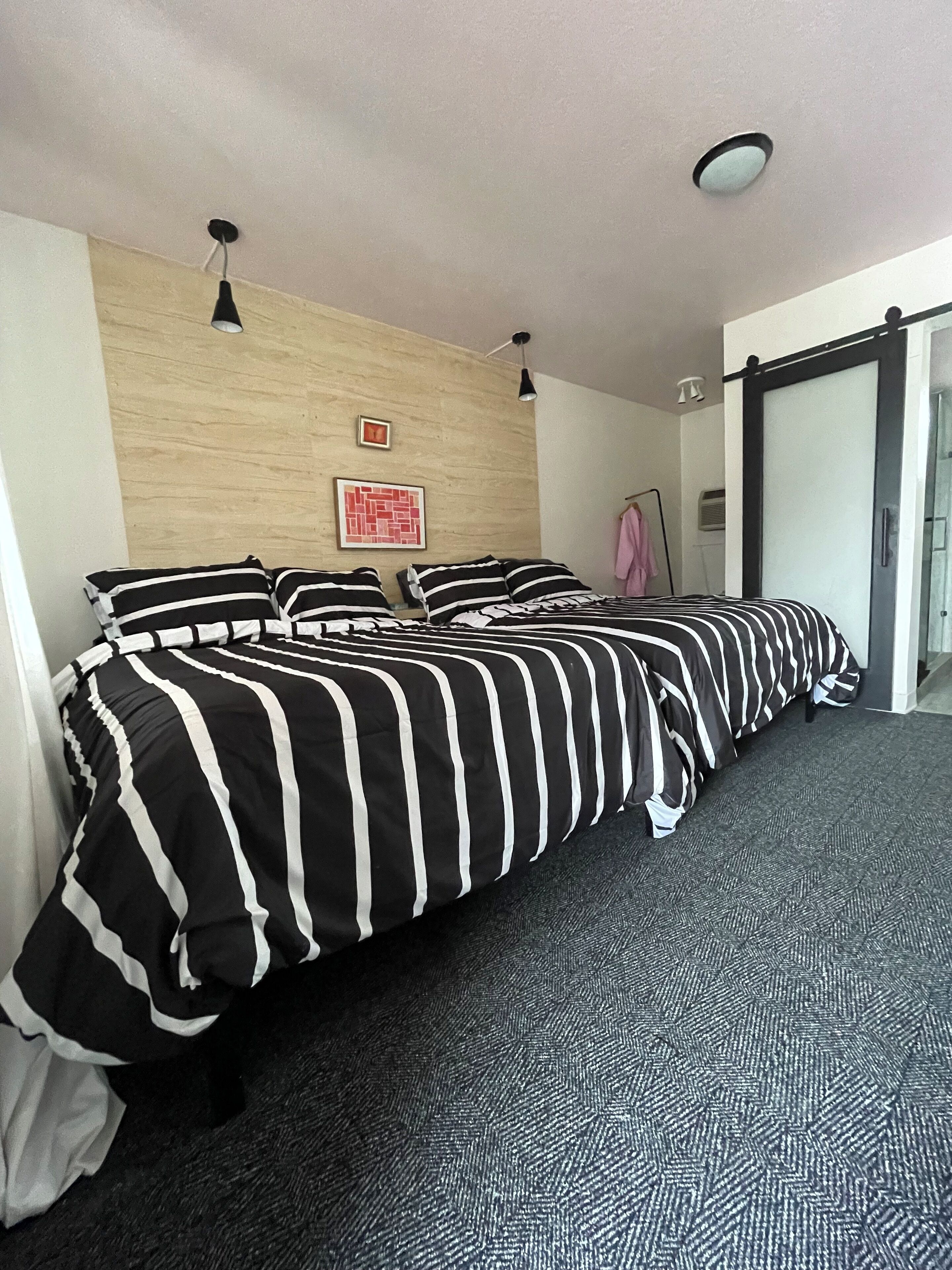 Habitación de hotel con cama, pared de madera, un cuadro, perchero y puerta corredera.