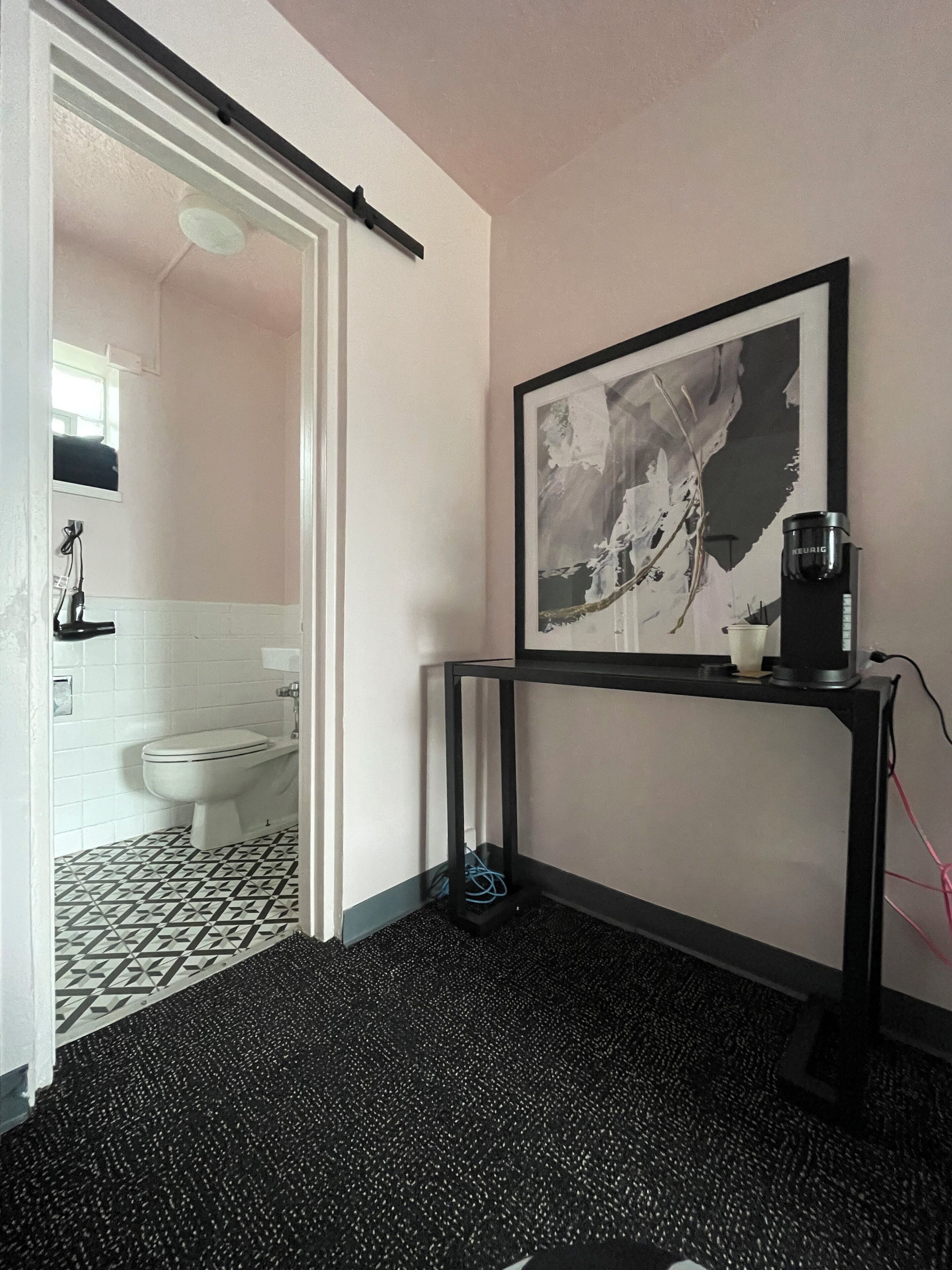 Un bagno con il water, un lavandino e una doccia. Si nota un quadro appeso al muro e una macchina da caffè su un tavolo.