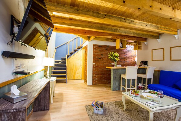 Un vaste espace de vie doté d’un plafond en bois, d’un canapé bleu, d’une table à manger où est dressé un repas, et d’un escalier menant à un niveau supérieur.