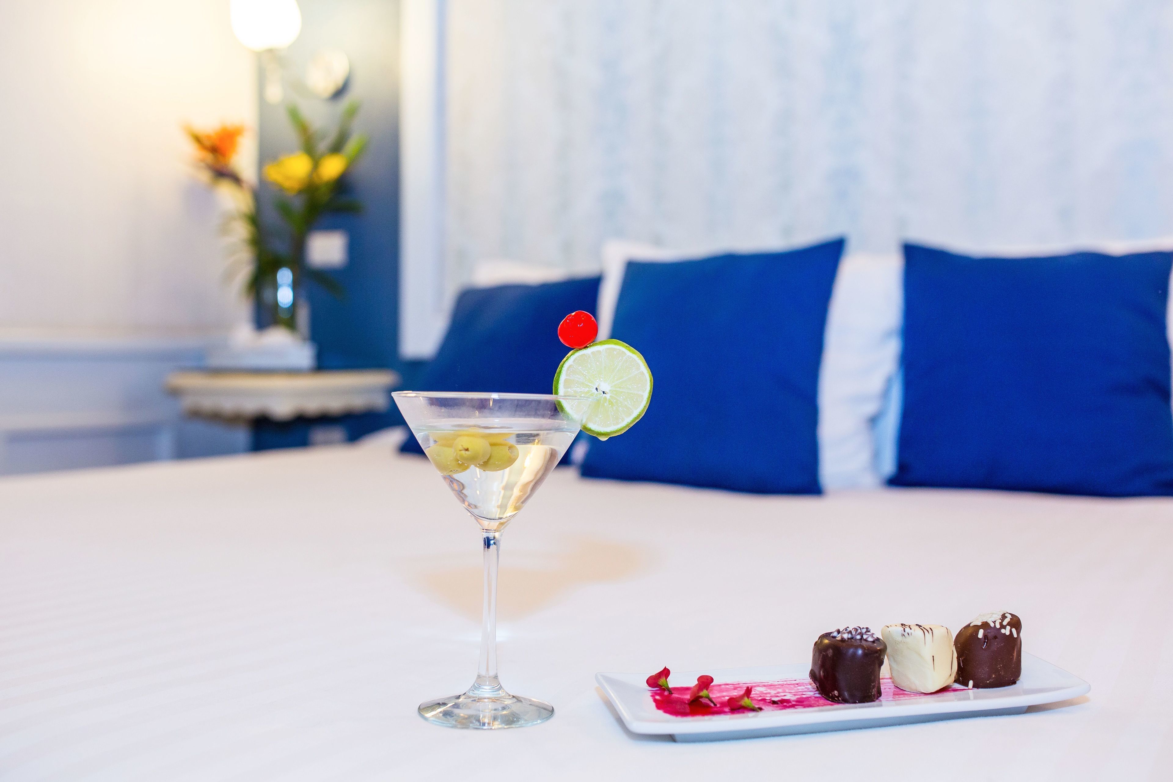 A martini glass with a lime slice and cherry on the rim, a plate with chocolate-covered desserts, and a bed with blue pillows in the background.