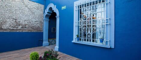Un patio con una pared azul, una ventana con rejas de metal blanco y un camino de ladrillo.