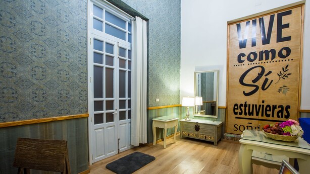 Une pièce avec un plancher en bois, une porte blanche, une petite table avec une lampe et un mur orné d’un panneau décoratif.