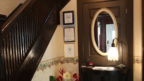 A staircase with dark wood railings, a wall-mounted mirror, a small vanity with a lamp, and a floral arrangement on the floor. - Ambiance by the Falls B&B (Niagara Falls)