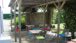 Un patio al aire libre con mesas y sillas, una pérgola de madera y una valla verde al fondo.