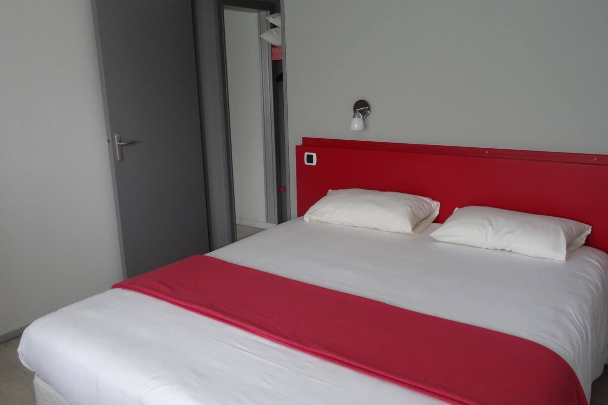 Una cama con ropa blanca y un cabecero rojo en una habitación con puerta gris y paredes blancas.