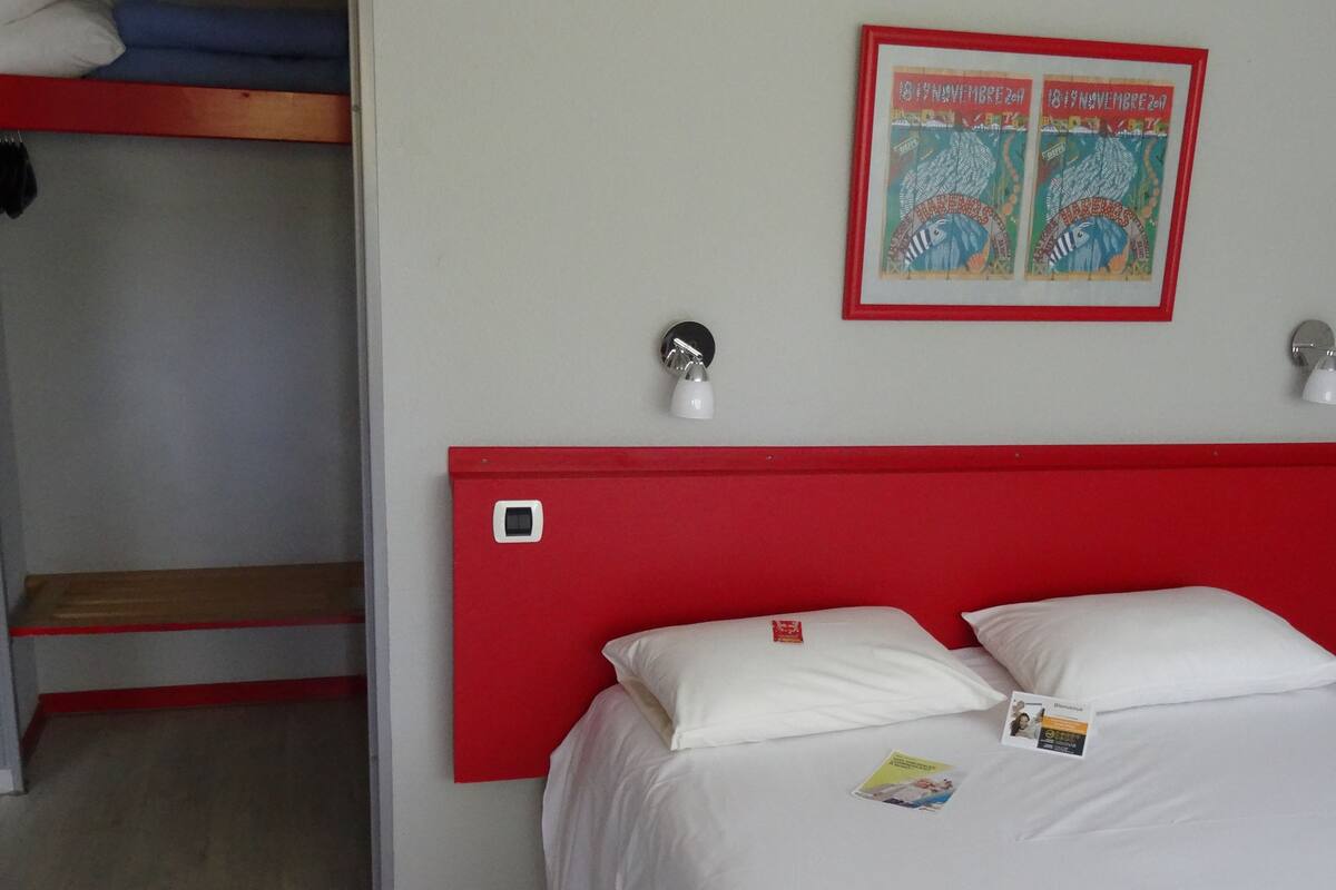 Una habitación de hotel con una cama litera, cabecera roja, dos lámparas de noche y un póster enmarcado en la pared.