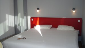 Habitación de hotel con una cama individual, ropa de cama blanca, cabecera roja, dos luces fijadas a la pared y una puerta a la izquierda.