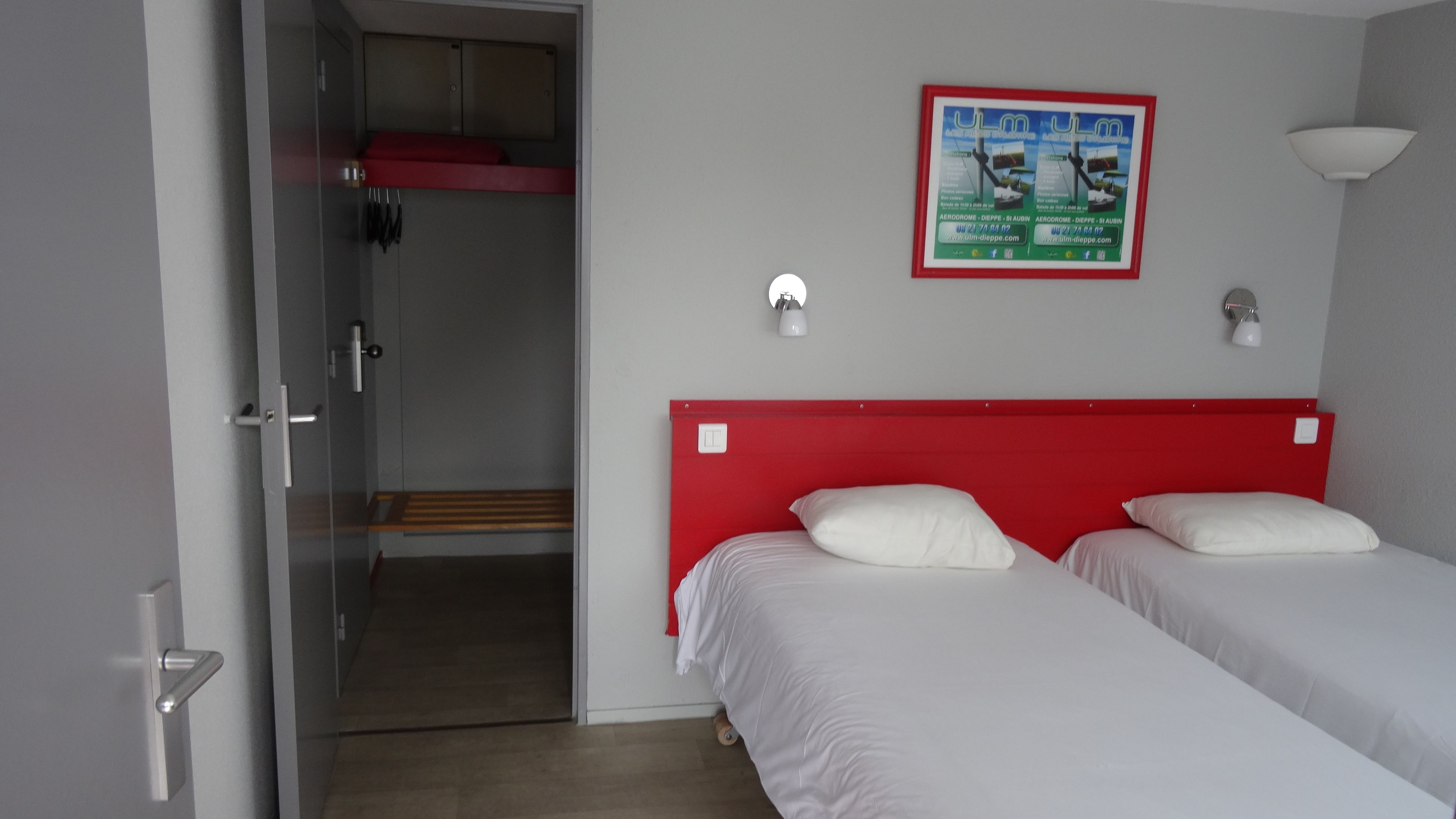 Habitación de hotel con dos camas individuales, un cabecero rojo, zona de ducha y un cartel en la pared.
