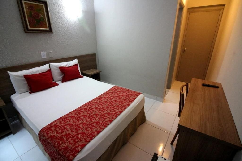 A hotel room with a bed, a wooden nightstand, a desk, and a door.