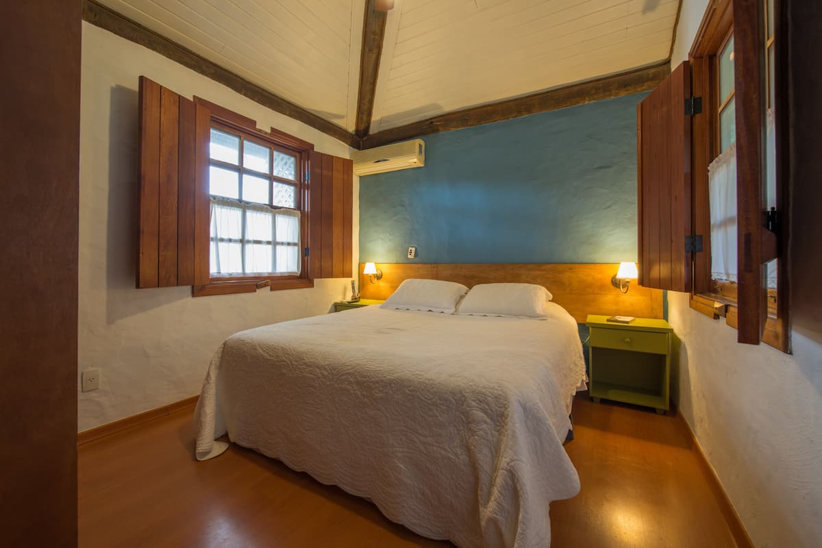 A bedroom with a large bed, wooden headboard, two bedside tables with lamps, a window with shutters, and an air conditioning unit on the wall.