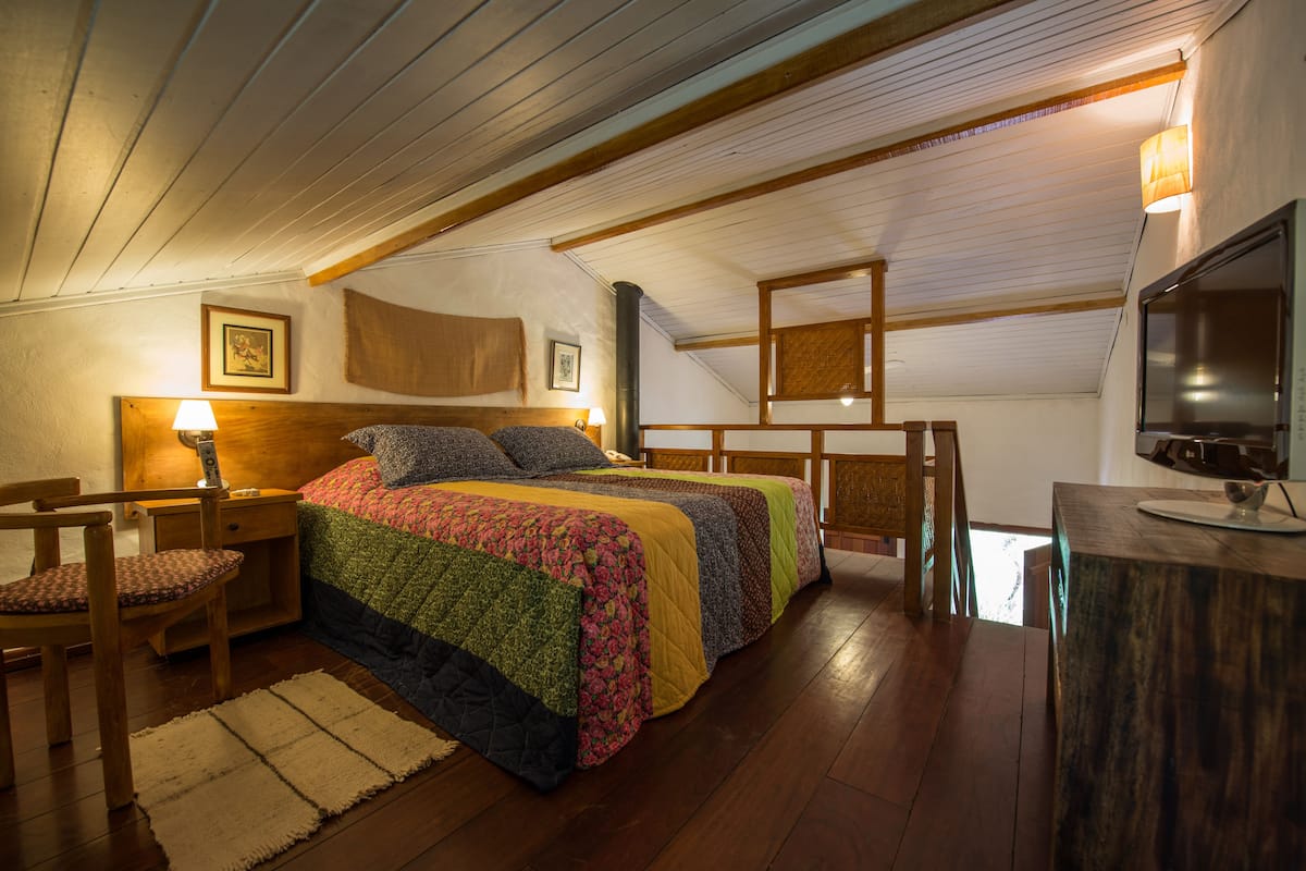 A cozy bedroom with a wooden ceiling, a bed with colorful bedding, a desk with a lamp, and a TV on a wooden stand.