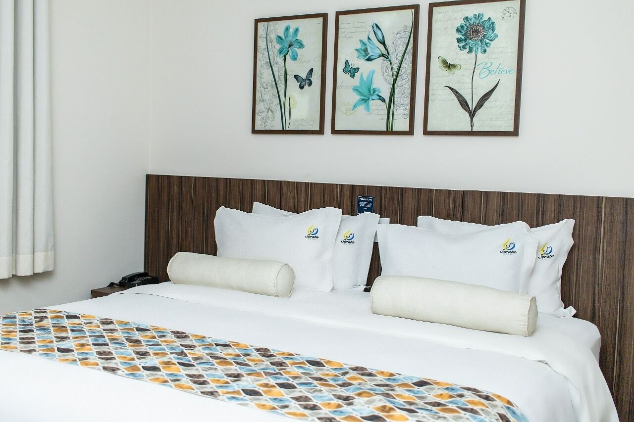 A hotel room with a neatly made bed, a wooden headboard, and three framed floral artworks on the wall.