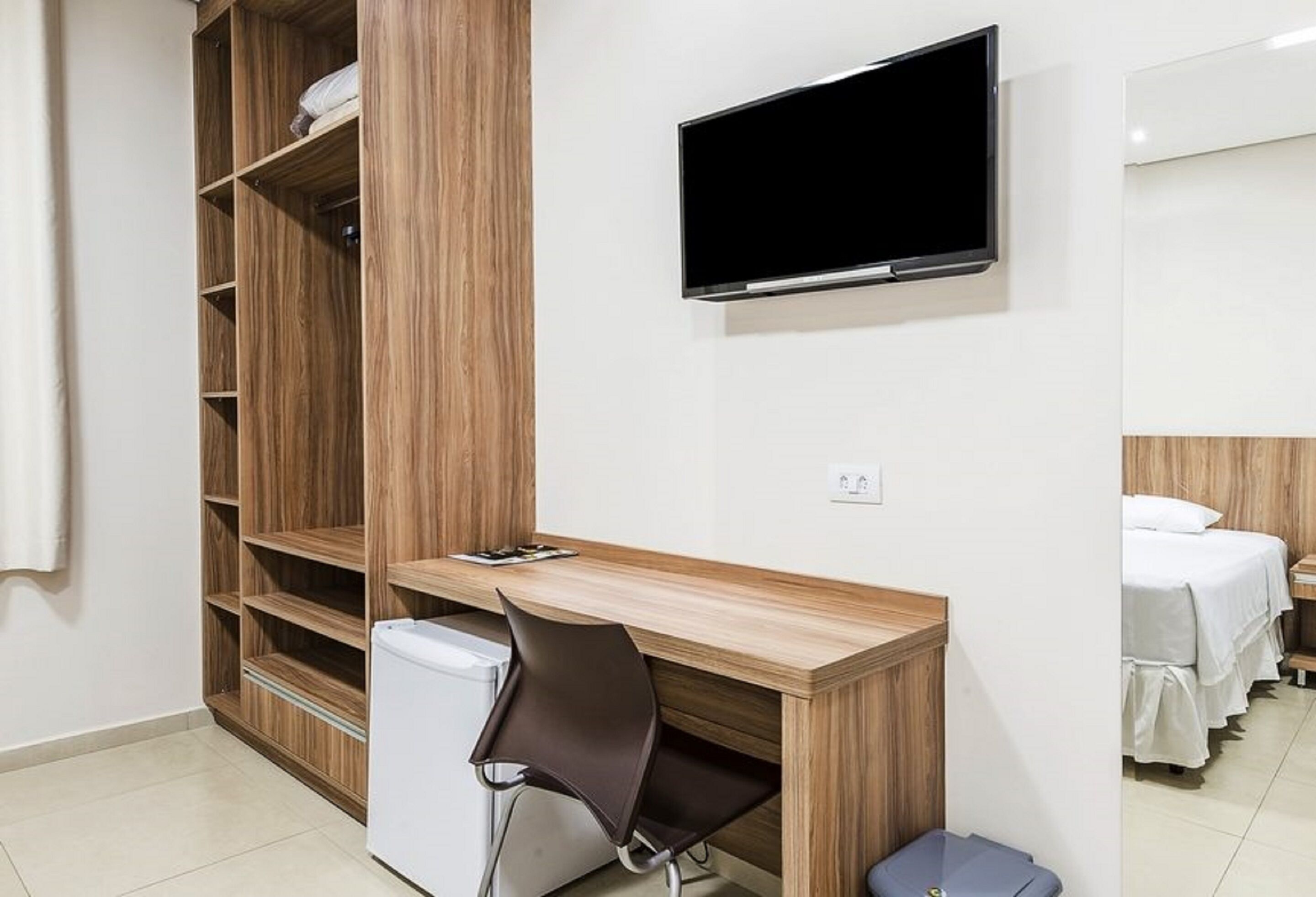 A compact room with a wooden desk, a chair, a television mounted on the wall, and a bed with white bedding.