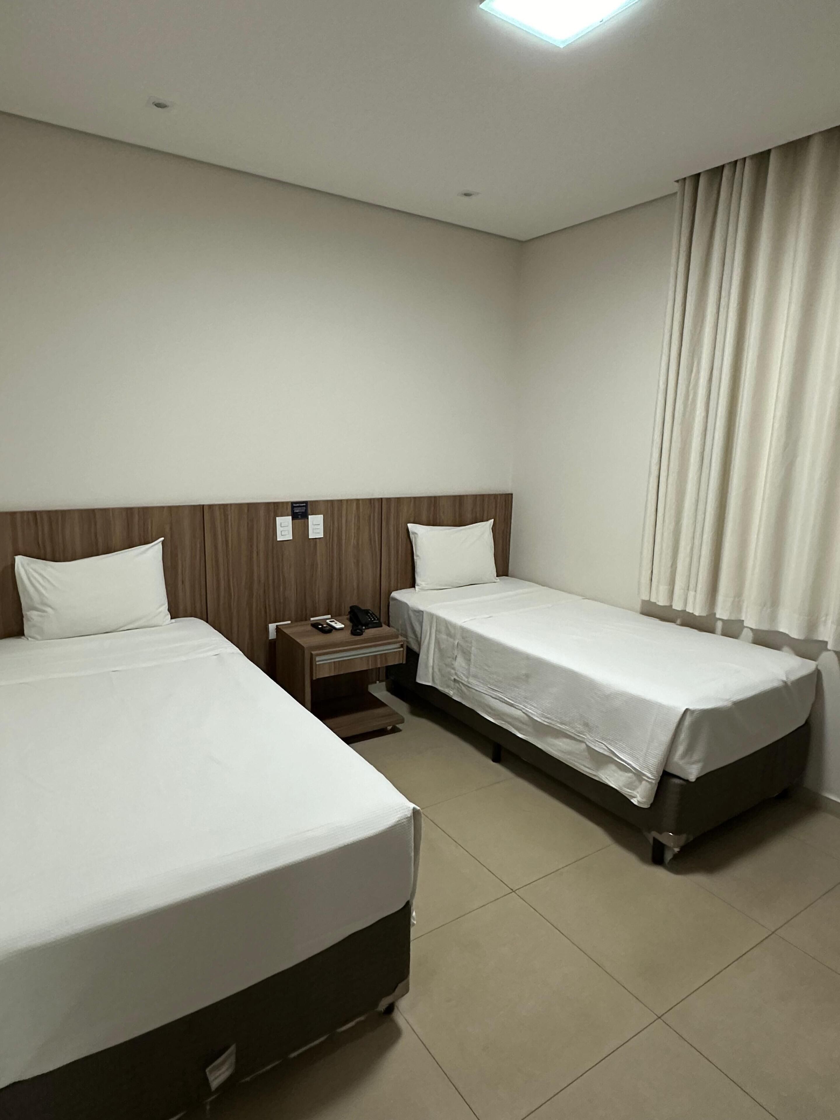 Habitación de hotel con dos camas, una mesita de noche con teléfono y control remoto, y una ventana con cortinas.