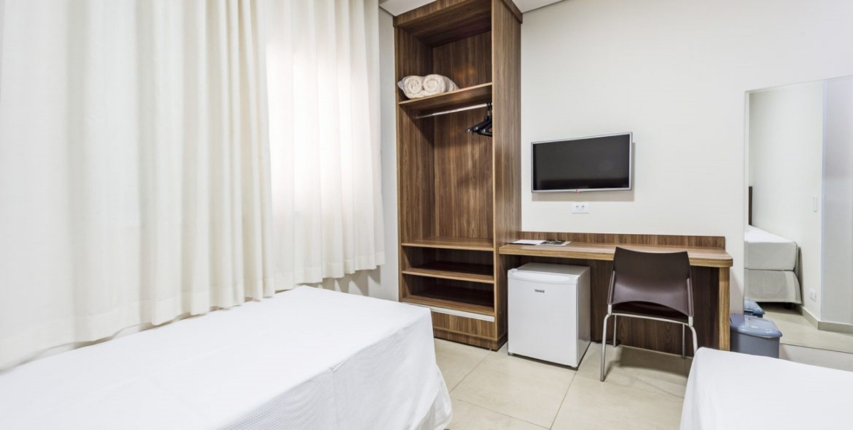 A hotel room with a bed, a desk with a chair, a television, a mini-fridge, and a wardrobe.