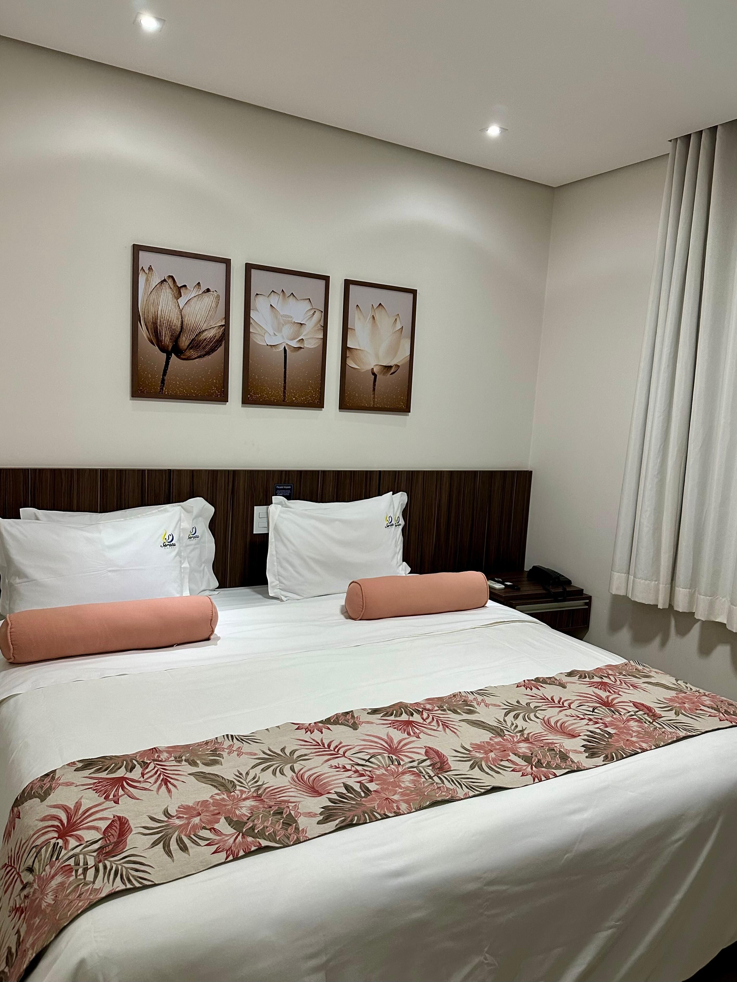 Quarto de hotel com cama bem arrumada, almofadas brancas, colcha florida e três quadros florais na parede.