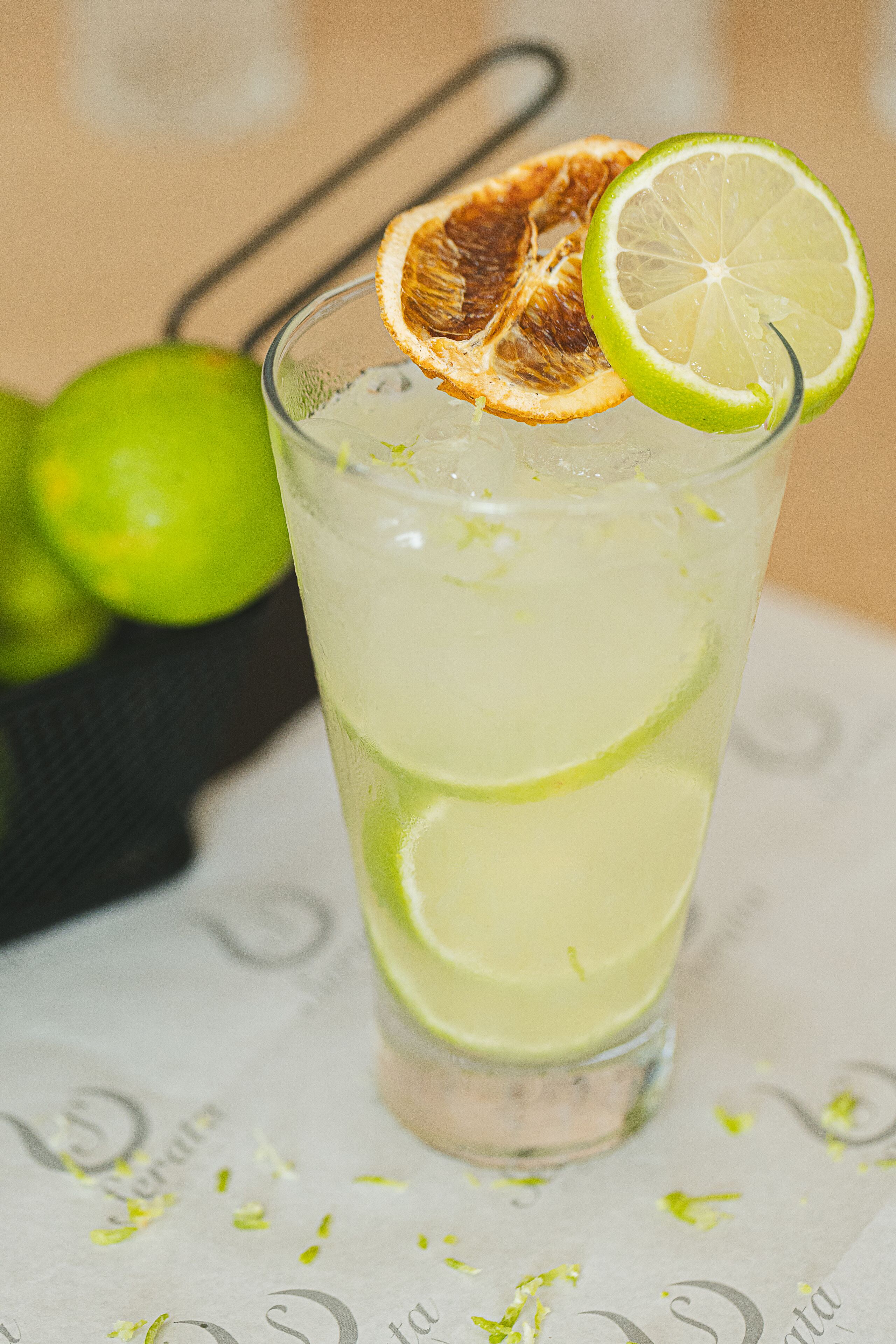 A tall glass with a lime slice and dried orange slice garnish, placed on a table with a menu underneath.