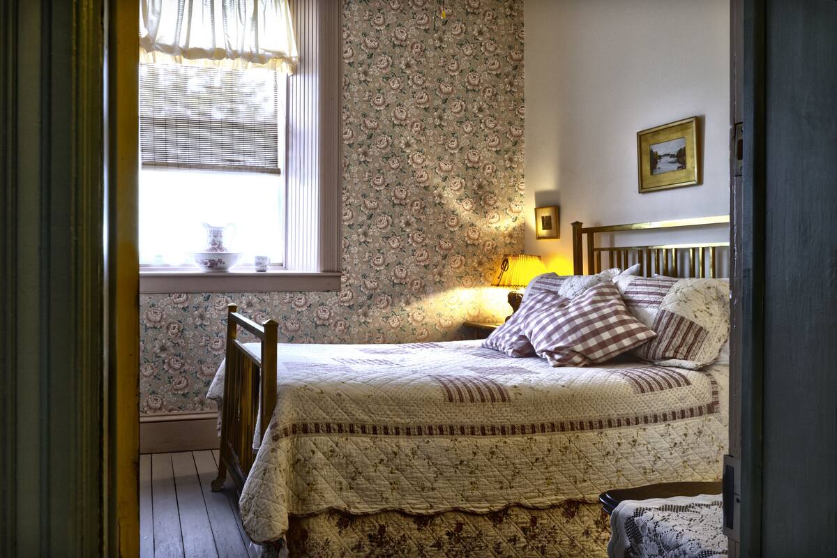 A bedroom with a wooden bed, floral wallpaper, a window with a curtain, and a bedside lamp.