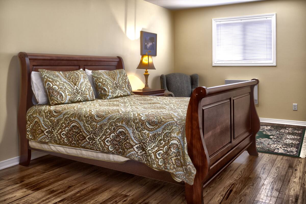 A bedroom with a wooden bed, patterned bedspread, bedside lamp, chair, and a window with blinds.
