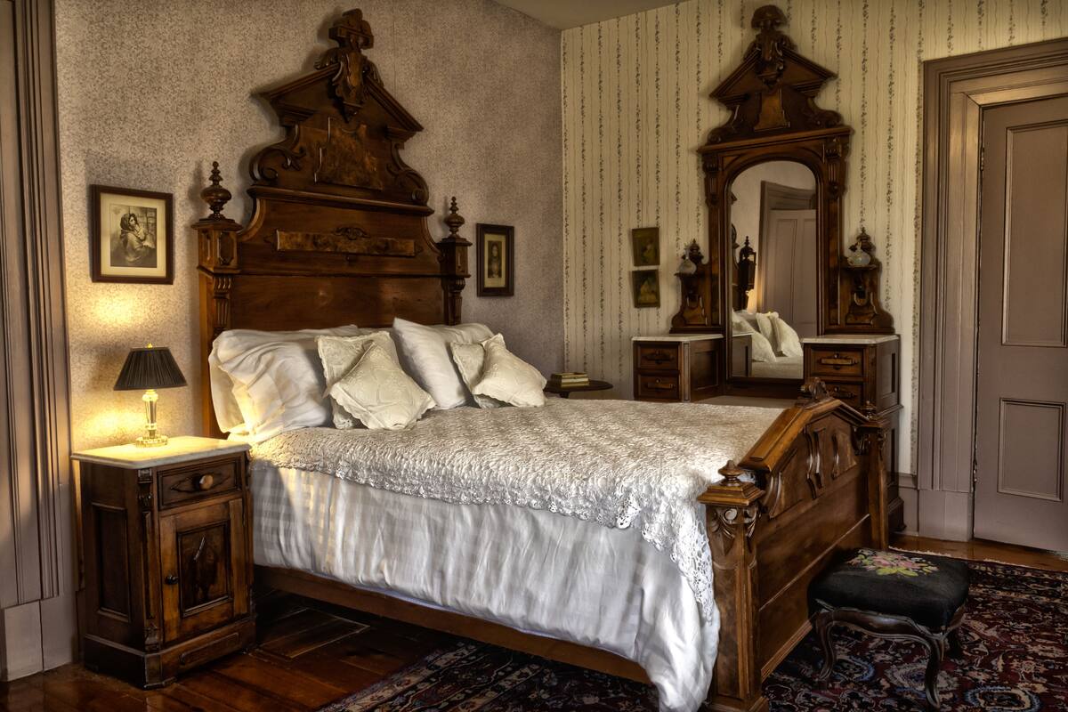 A traditional bedroom with a wooden four-poster bed, a nightstand with a lamp, a dresser, and a mirror.