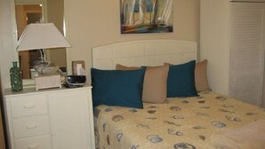 A bedroom with a bed, a bedside table with a lamp, a dresser, and a painting on the wall. - Sea Whale Cottages (Middletown)