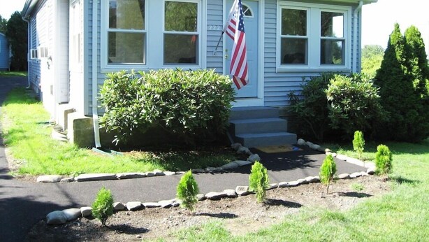 A single-story house with a front porch, a flag, and a well-maintained garden with small shrubs and a stone border. - Sea Whale Cottages (Middletown)