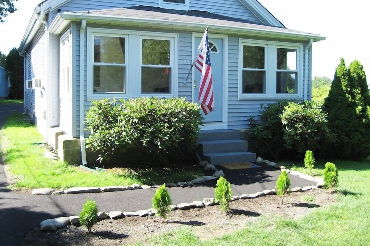 A single-story house with a front porch, a flag, and a well-maintained garden with small shrubs and a stone border.
