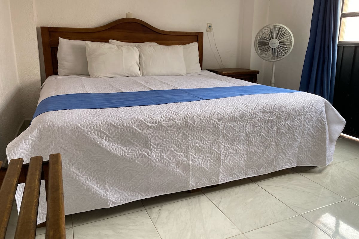 A neatly made bed with a white quilt and a blue bedspread, a wooden headboard, a bedside table with a fan, and a window with blue curtains.