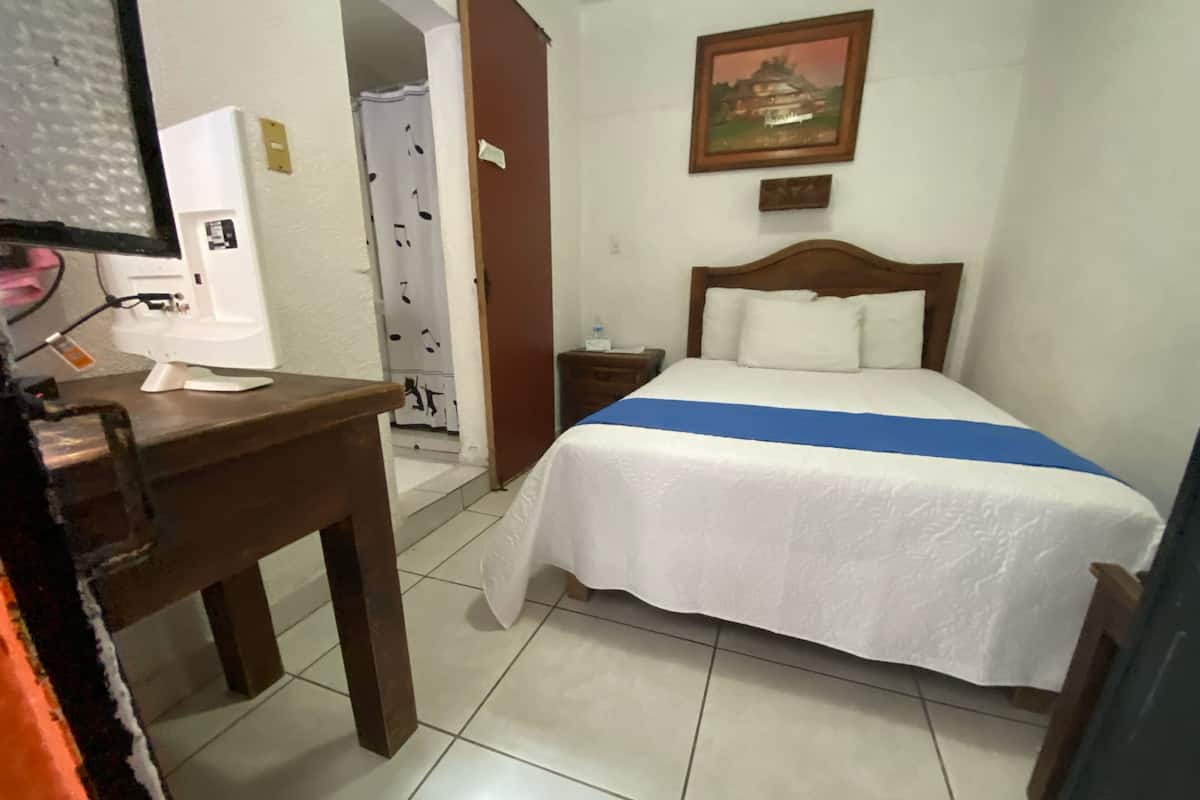 A hotel room with a bed, a desk, a nightstand, a painting on the wall, and a bathroom visible through an open door.